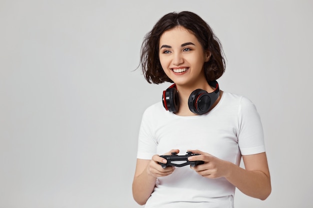 Bella ragazza coinvolta riccia che gioca ai videogiochi sulla console su sfondo grigio