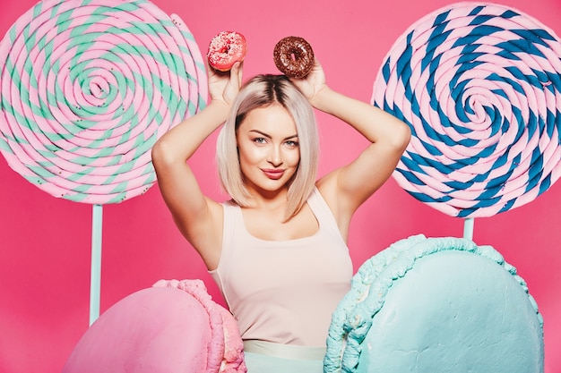 Bella ragazza con capelli biondi che indossa top e berretto rosa in piedi con enormi dolci lecca-lecca in studio rosa