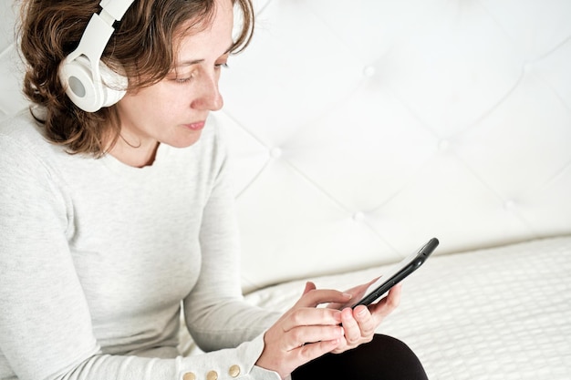 Bella ragazza con capelli castano chiaro che si siede sul letto indossando le cuffie guardando il suo smartphone