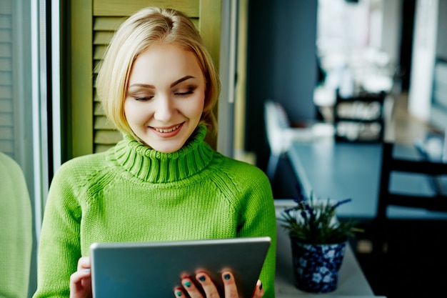 Bella ragazza con capelli chiari che indossa un maglione verde seduto nella caffetteria con tablet, concetto di freelance, shopping online, ritratto.