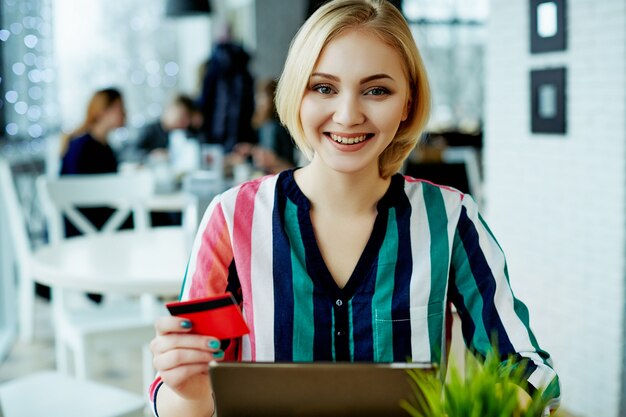 Bella ragazza con capelli chiari che indossa una camicia colorata che si siede nella caffetteria con tablet, carta di credito e tazza di caffè, concetto di freelance, shopping online.