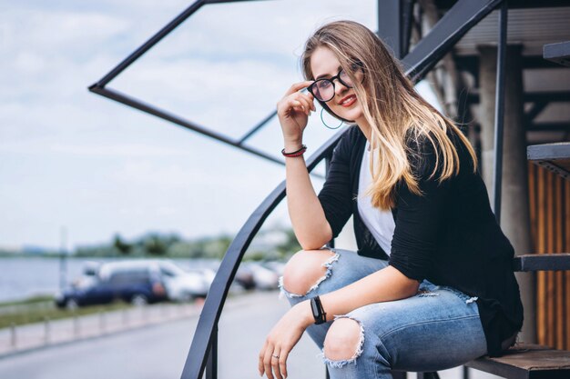 Bella ragazza con capelli lunghi e vetri che si siedono sulle scale del metallo sui precedenti di legno della casa con i bordi verticali. Donna che sorride e che esamina macchina fotografica