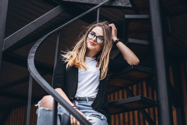 Bella ragazza con capelli lunghi e vetri che si siedono sulle scale del metallo sui precedenti di legno della casa con i bordi verticali