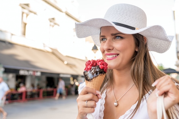 Bella ragazza con cappello bianco e borse per la spesa che mangia il gelato sulla strada della città