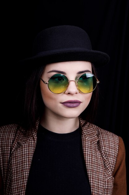 Bella ragazza con cappello e occhiali rotondi sta posando in studio su sfondo nero