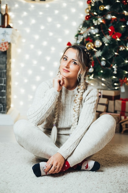 Bella ragazza con due trecce sullo sfondo di un albero di Natale e un caminetto in attesa di Natale e Capodanno, carina