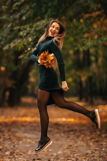 Bella ragazza con foglie d'autunno in un parco che salta e sorride