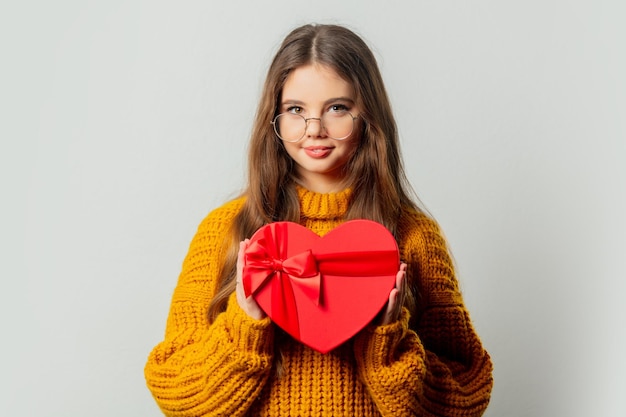 Bella ragazza con gli occhiali e maglione giallo con scatola regalo a forma di cuore su sfondo bianco