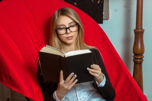 Bella ragazza con gli occhiali seduto su una comoda sedia rossa con un libro nero tra le mani e legge