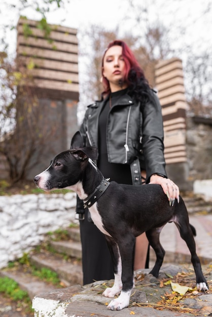 Bella ragazza con i capelli bordeaux e in un abito elegante con un simpatico cucciolo al guinzaglio In posa nel parco Dog mix Staffordshire Terrier e Pit Bull Terrier