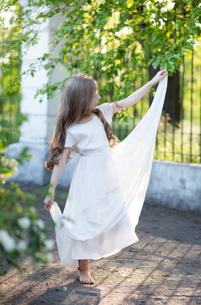 bella ragazza con i capelli lunghi biondi in un abito di seta bianca delicata danza balletto a piedi nudi in un parco con una bella luce