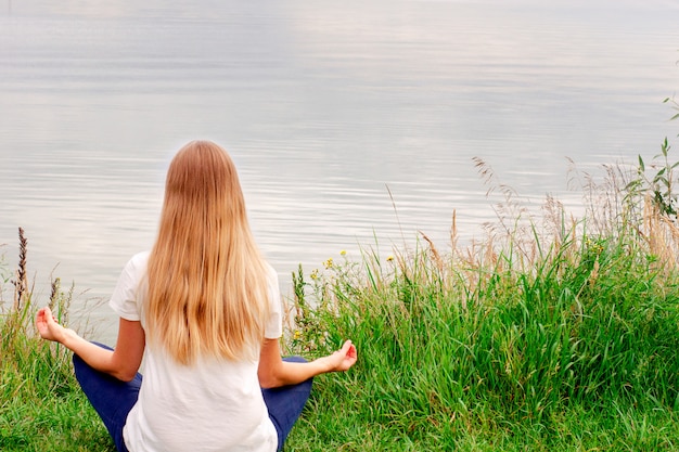 Bella ragazza con i capelli lunghi è seduto sulla riva. La vista dal retro. Tramonto. Pace e tranquillità Yoga in riva al lago