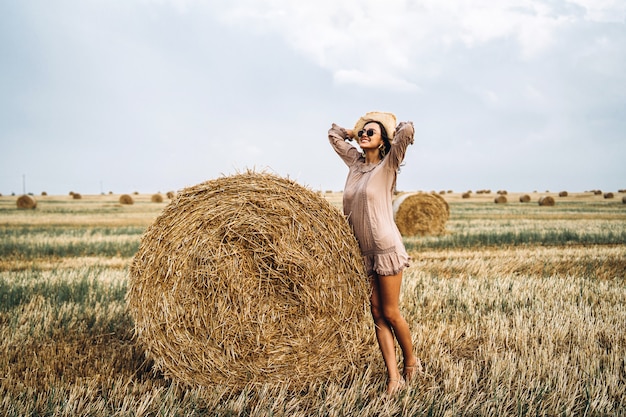 Bella ragazza con i capelli lunghi in occhiali da sole e cappello di paglia in posa su un campo di grano vicino a balle di fieno. Felice bruna in abito estivo