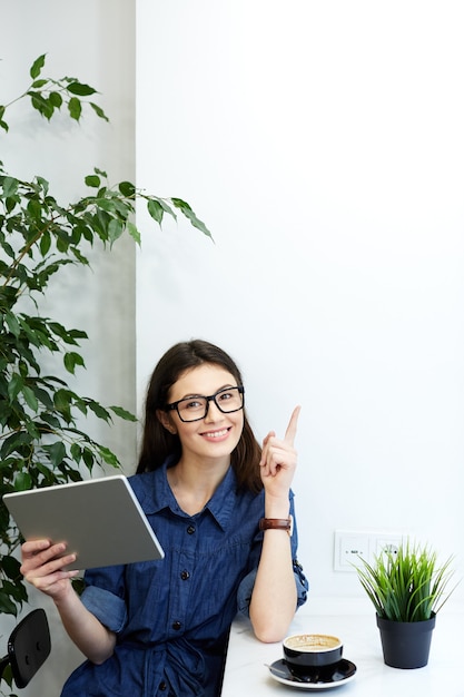 Bella ragazza con i capelli neri che indossa camicia blu spogliata e occhiali seduti nella caffetteria con tavoletta e tazza di caffè, concetto di freelance, ritratto, avendo una buona idea.