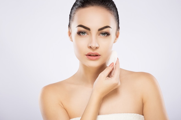 Bella ragazza con i capelli neri fissati dietro, grandi occhi, sopracciglia folte e spalle nude in piedi al muro grigio e che tiene una spugna per la pulizia cosmetica.