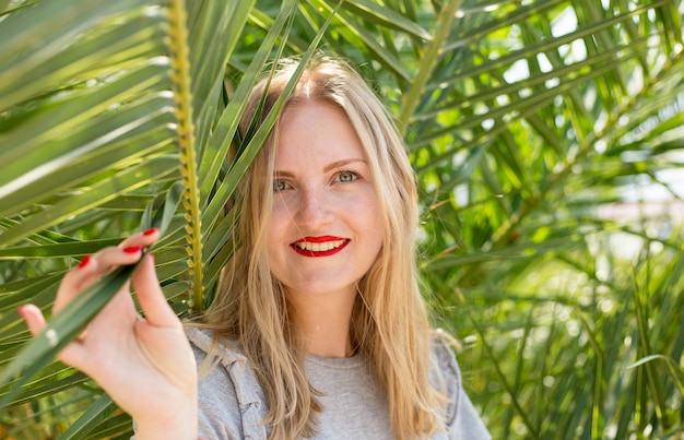 Bella ragazza con le labbra rosse che sorride e che posa nella foresta tropicale. Ritratto di donna tenera che guarda fuori dalla foglia di palma