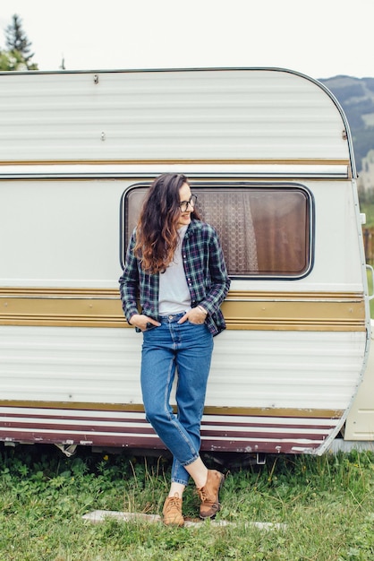 Bella ragazza con lo zaino in viaggio Ragazza in piedi vicino al rimorchio che viaggia in montagna Donna con gli occhiali che viaggia in auto Ragazza viaggiatore thumbs up