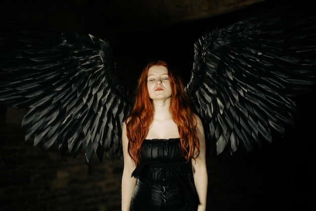 Bella ragazza con lunghi capelli rossi e ali nere in una stanza buia