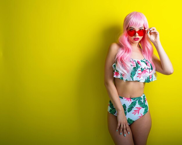 Bella ragazza con parrucca rosa e bikini colorato in posa su sfondo giallo Donna con capelli lunghi artificiali e occhiali rosa a forma di cuore