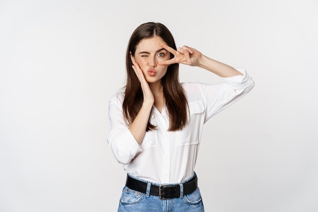 Bella ragazza con pelle sana, che mostra il segno di pace sul viso e strizza l'occhio, in piedi su sfondo bianco. Concetto di persone ed emozioni.