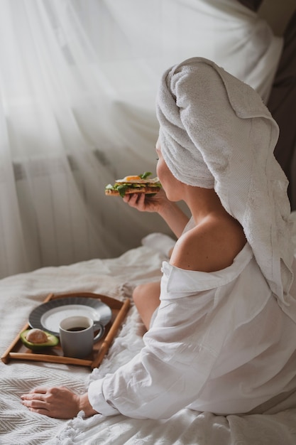 Bella ragazza con un asciugamano bianco in testa si siede a letto e fa colazione con un panino sano con caffè