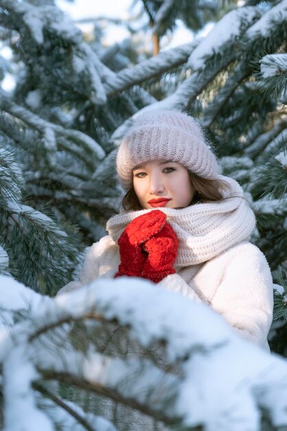 Bella ragazza con un cappello caldo e guanti si erge tra i pini innevati. Giornata invernale soleggiata.