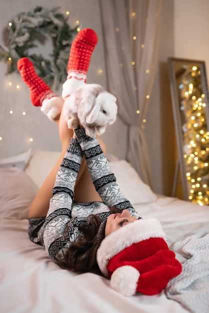 Bella ragazza con un maglione natalizio e un cappello rosso da Babbo Natale giace sul letto e tiene in mano un piccolo coniglio bianco Celebrazione del Natale di capodanno