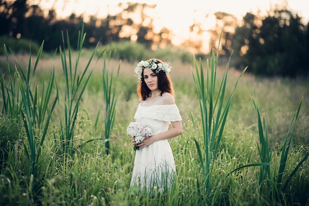 Bella ragazza con un mazzo di fiori e una corona in un vestito bianco