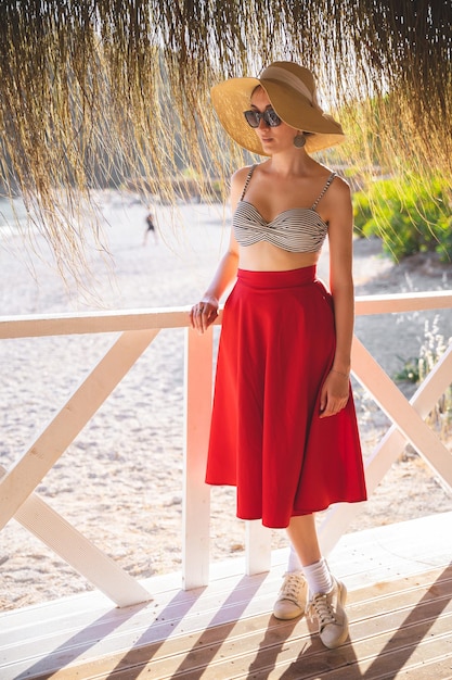 Bella ragazza con una gonna rossa e un cappello in estate su una terrazza sulla riva del mare