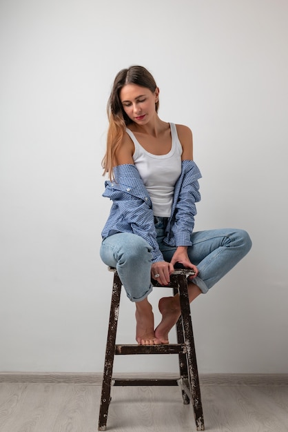 Bella ragazza con una maglietta bianca, jeans blu seduta su una sedia, sorridente e in posa