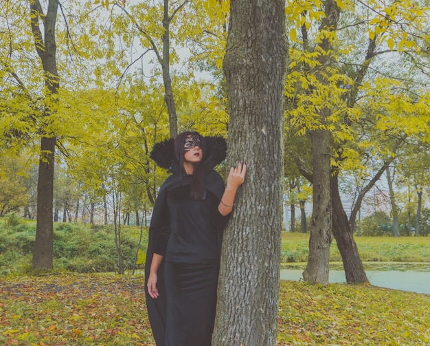 Bella ragazza con una maschera nera e un abito è appoggiata all'albero.