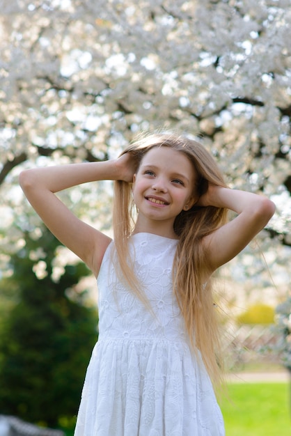 Bella ragazza dagli occhi azzurri con lunghi capelli biondi in un abito bianco che cammina nel giardino fiorito