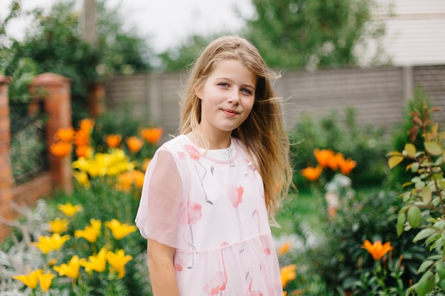 bella ragazza dagli occhi blu con lunghi capelli biondi. bambina in un vestito di fenicottero rosa. ragazza nel giardino fiorito. estate luminosa, foto emotiva. giardino fiorito grande, spesso e luminoso vicino alla casa.