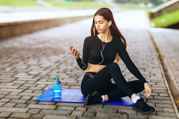 Bella ragazza dai capelli castani ascolta musica mentre è seduto su un tappetino da ginnastica e si riposa dopo aver praticato sport