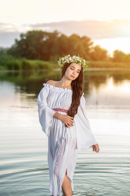 Bella ragazza dai capelli nera in abito vintage bianco e corona di fiori in piedi nell'acqua del lago. Bagliore di sole.