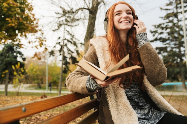 Bella ragazza dai capelli rossi che ascolta la musica con le cuffie mentre è seduto su una panchina, leggendo un libro