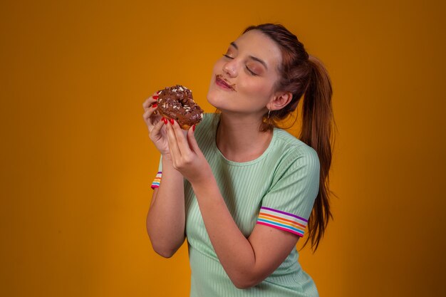 Bella ragazza dai capelli rossi che mangia ciambelle al cioccolato su sfondo giallo. ragazza che mangia ciambelle