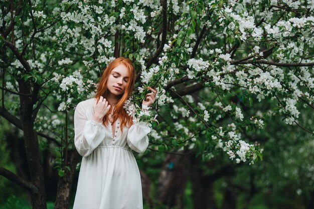 Bella ragazza dai capelli rossi in un abito bianco tra meli in fiore nel giardino