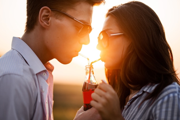 Bella ragazza dai capelli scuri e un giovane uomo in occhiali da sole stanno bevendo da una bottiglia un drink attraverso le cannucce all'aperto in una giornata di sole. .