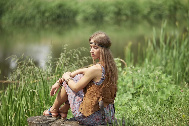 Bella ragazza del hippie che si siede su un ceppo vicino allo stagno