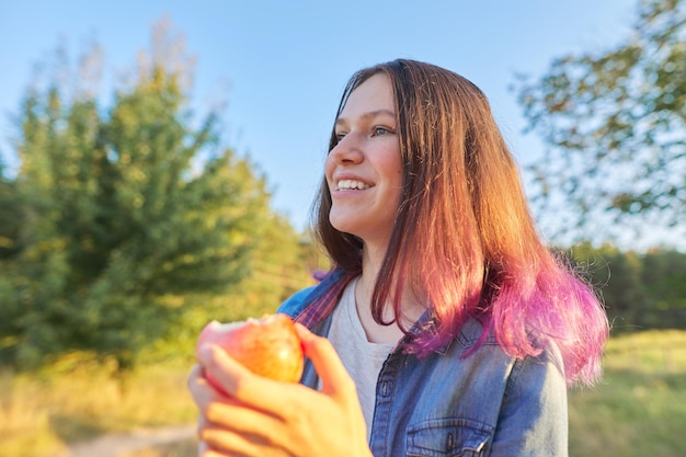 Bella ragazza dell'adolescente che mangia mela rossa saporita succosa matura all'aperto. Bellissimo paesaggio naturale sullo sfondo, ora d'oro. Cibo sano naturale