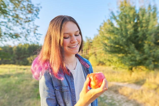 Bella ragazza dell'adolescente che mangia mela rossa saporita succosa matura all'aperto. Bellissimo sfondo del paesaggio naturale, ora d'oro. Cibo sano naturale