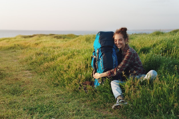 Bella ragazza della viandante che abbraccia il suo zaino nell&#39;erba e nel sorridere