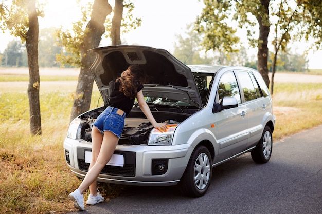 Bella ragazza dello studente che indossa i mini pantaloncini e la maglietta nera che si appoggia sul cofano aperto dell'automobile di un'automobile moderna ripartita