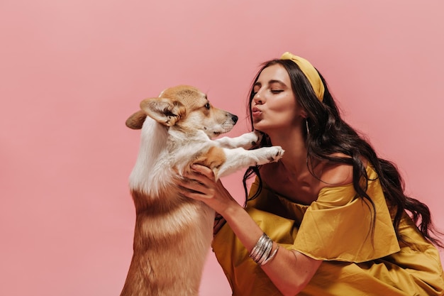 Bella ragazza di buon umore con capelli castani in bandana gialla e vestiti alla moda in posa con un cane carino su sfondo isolato