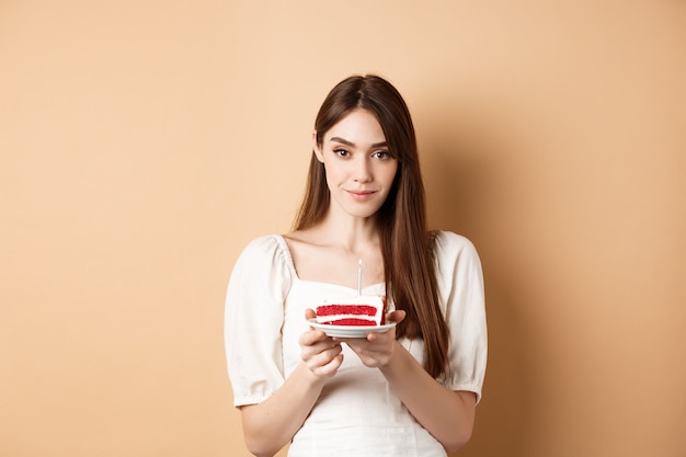 Bella ragazza di compleanno che tiene la torta con la candela, esprimendo il desiderio, celebrando il b-day su fondo beige.