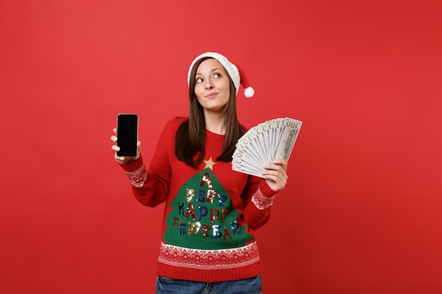 Bella ragazza di Santa che cerca tenendo un sacco di banconote in dollari, denaro contante telefono cellulare con schermo vuoto vuoto isolato su sfondo rosso. Felice Anno Nuovo 2019 celebrazione festa concetto.