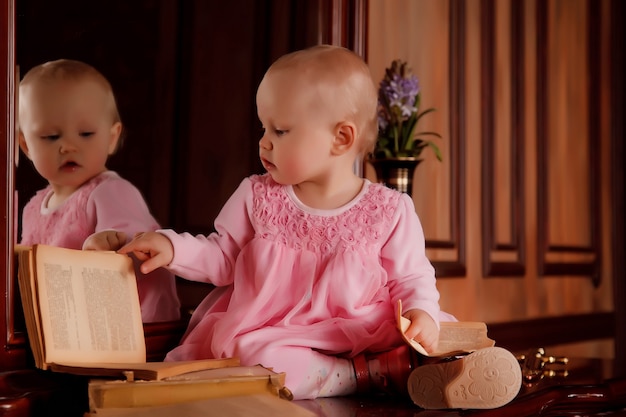 Bella ragazza di un anno in abito rosa con un vecchio libro è seduta sulla credenza vicino allo specchio