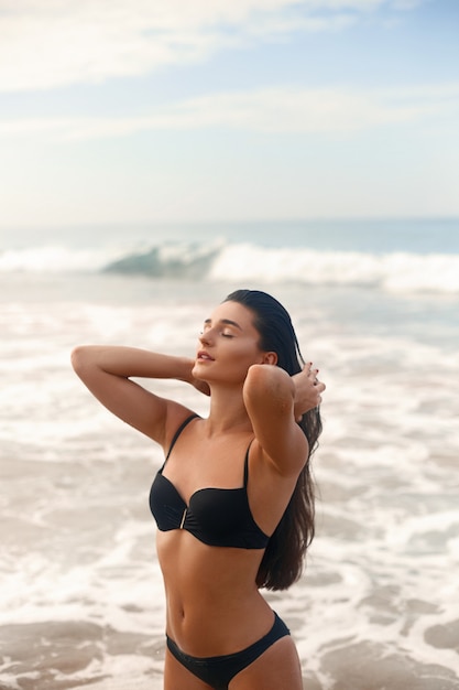 Bella ragazza esile di lusso in un bikini nero sulla spiaggia dell'oceano. Donna di bellezza con corpo abbronzato sexy, pancia piatta, figura perfetta. Riposa su un'isola tropicale.