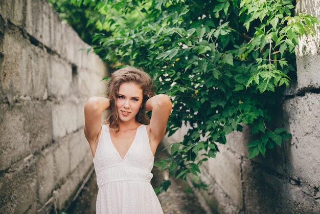 Bella ragazza felice con capelli naturali ricci in vestito bianco vicino alle foglie verdi dell'albero.